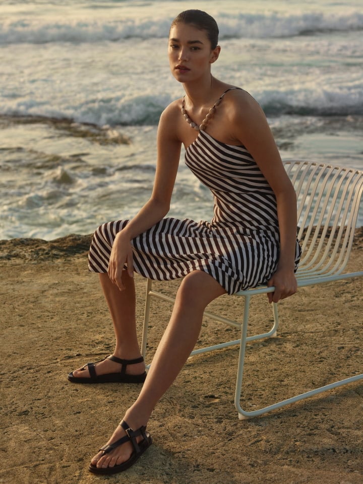A woman in a striped dress sits on a white chair on a stone surface in front of the ocean during daytime. She wears sandals.
