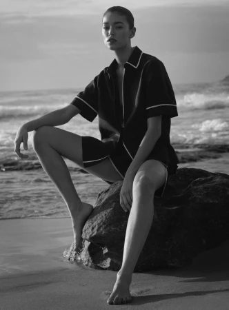 Black and white shot of a woman in a short suit sitting on a rock at the beach with the ocean and sky in the background.