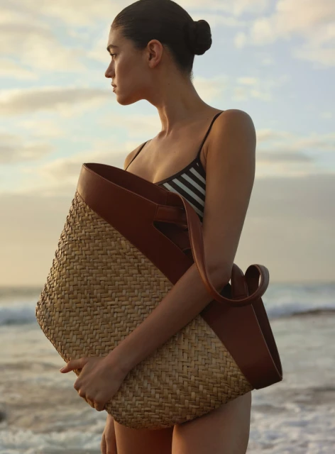 Woman in swimsuit holds a woven bag on a beach at sunset. The image focuses on the woman and the bag against an ocean background.