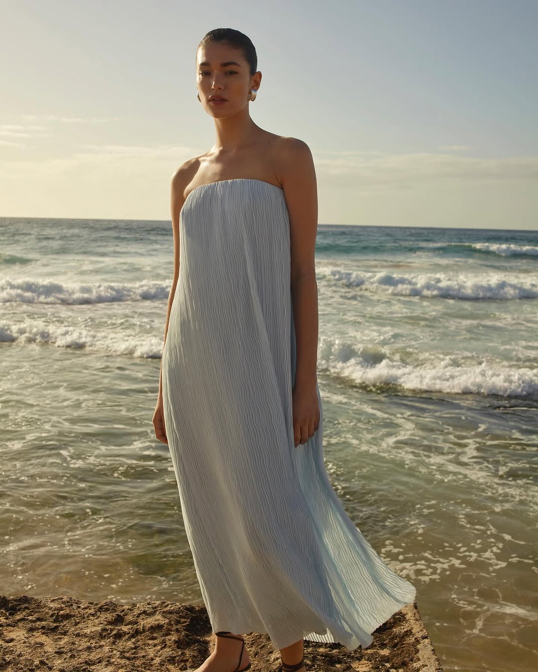 A woman in a light blue, strapless dress stands on a beach with the ocean behind her, against a clear sky and wearing minimal jewelry.
