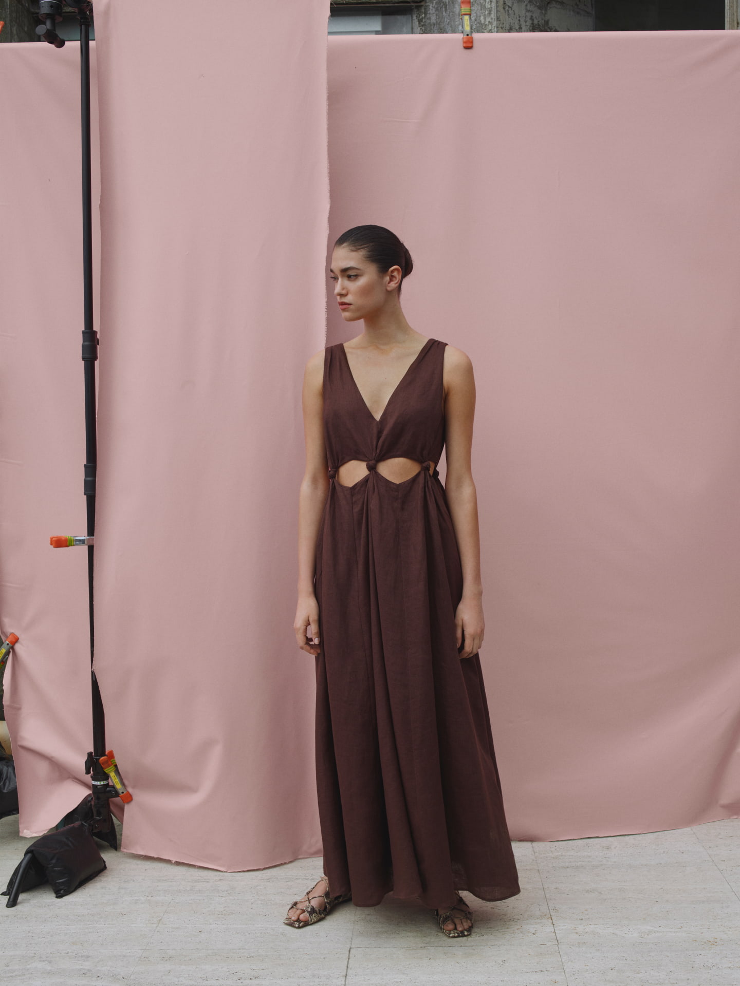 A slender woman in a brown V-neck dress with cutouts poses against a pink backdrop in a fashion photo, highlighted by soft lighting.