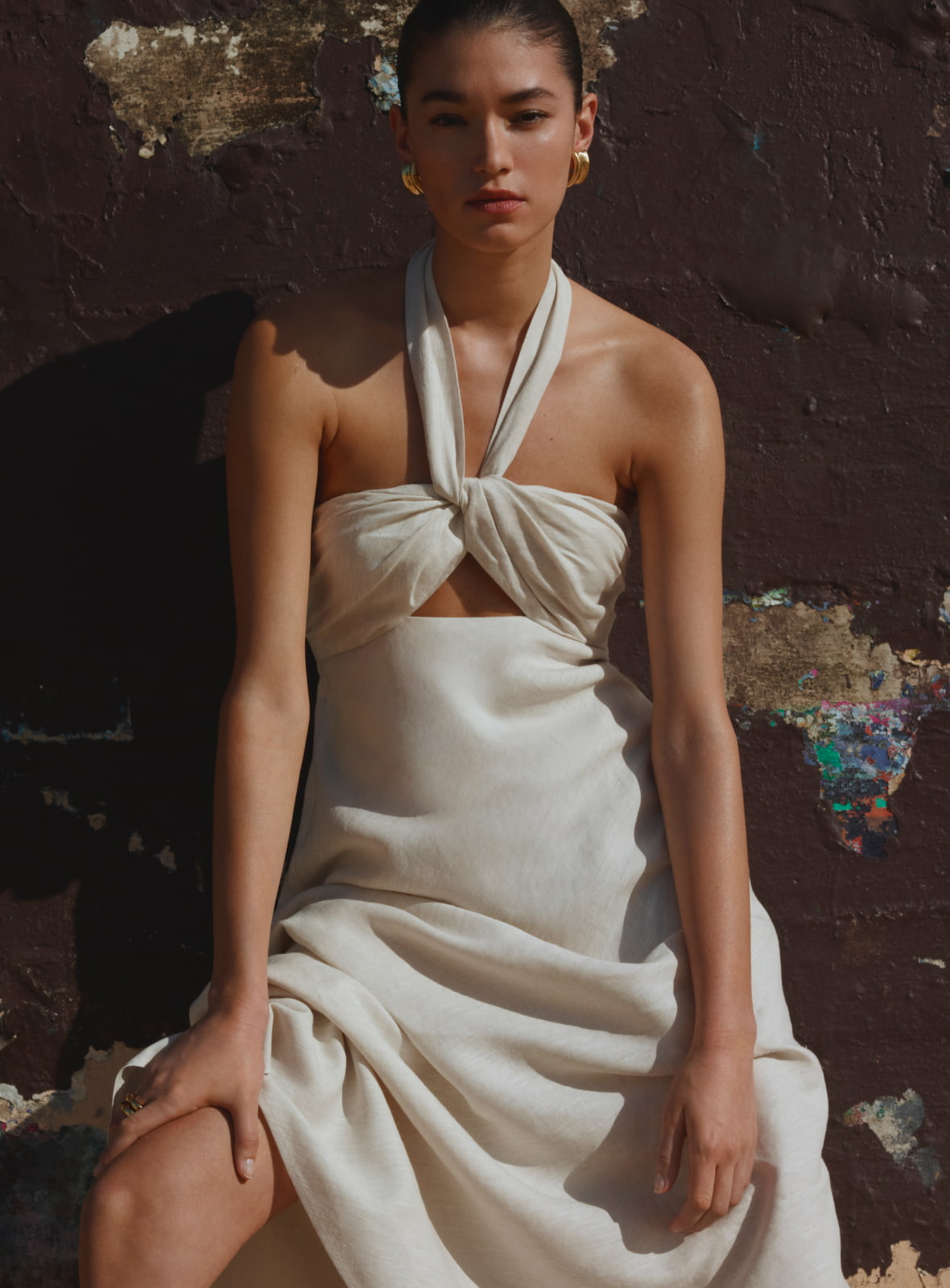 A woman in a cream-colored dress poses in front of a textured wall, showcasing fashion and style with elegant earrings.