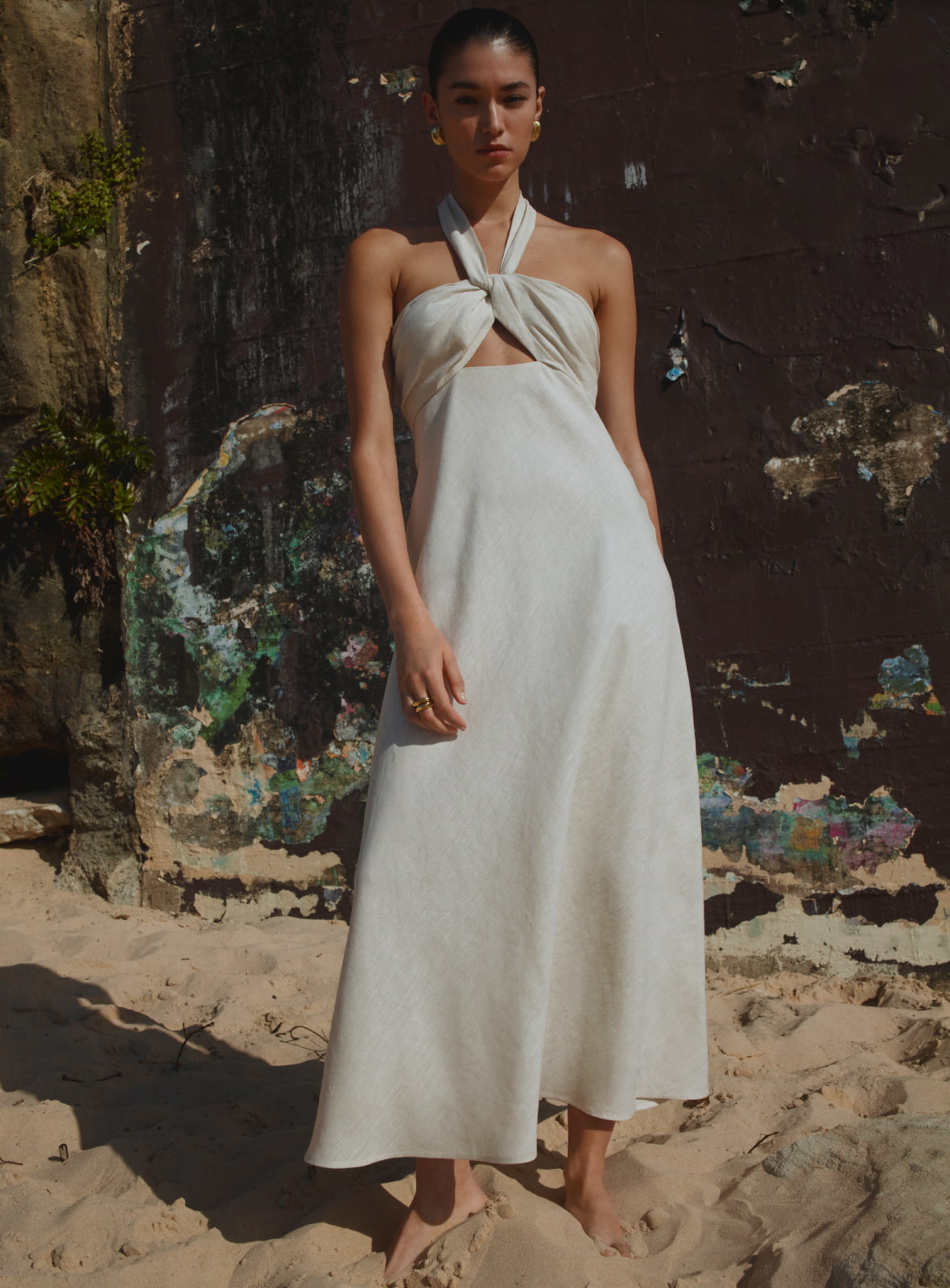 Model wearing a white halter-neck dress standing barefoot on a sandy beach in front of a textured rock wall.