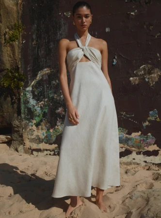 Model wearing a white halter-neck dress standing barefoot on a sandy beach in front of a textured rock wall.