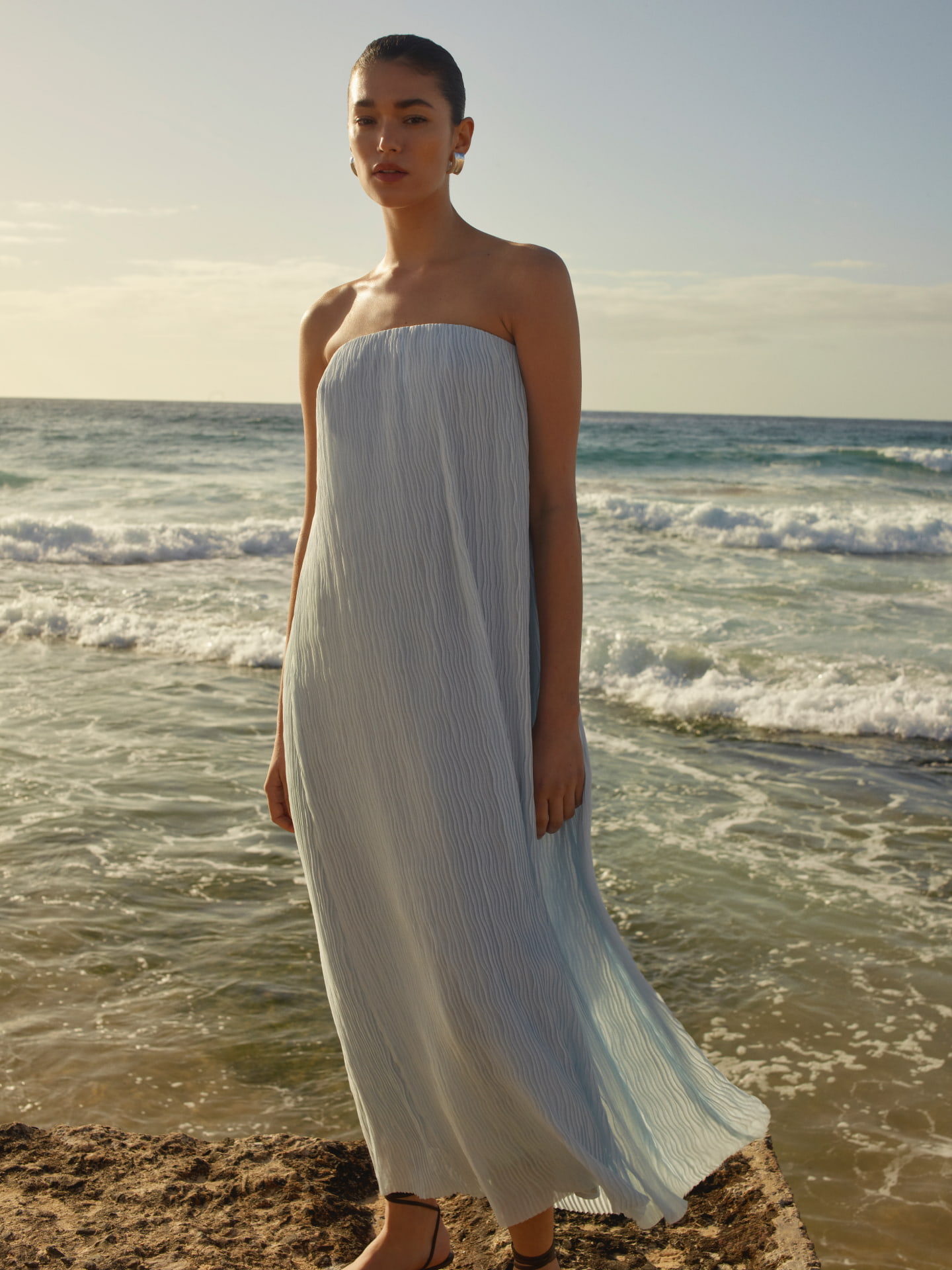 A woman in a light blue strapless dress stands on a beach with waves, sand, and a clear sky in a fashion or beauty shoot.