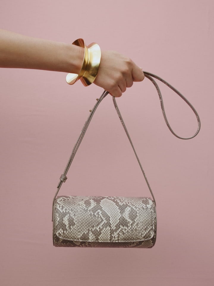 An arm holds a cylindrical snakeskin purse on a pink background, accessorized with a large, modern gold cuff bracelet.