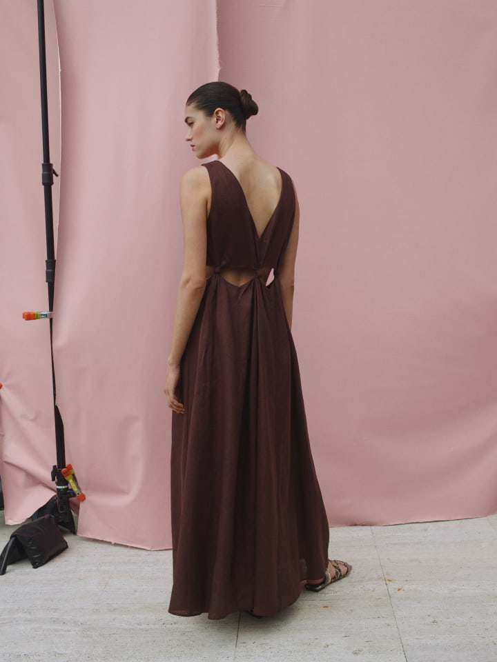 Woman in a sleeveless brown maxi dress poses in front of a pink backdrop in a studio, showcasing the dress's cut-out back and design.