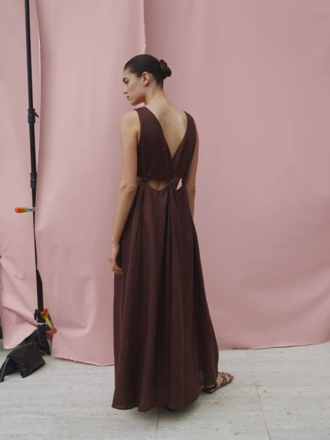 Woman in a sleeveless brown maxi dress poses in front of a pink backdrop in a studio, showcasing the dress's cut-out back and design.