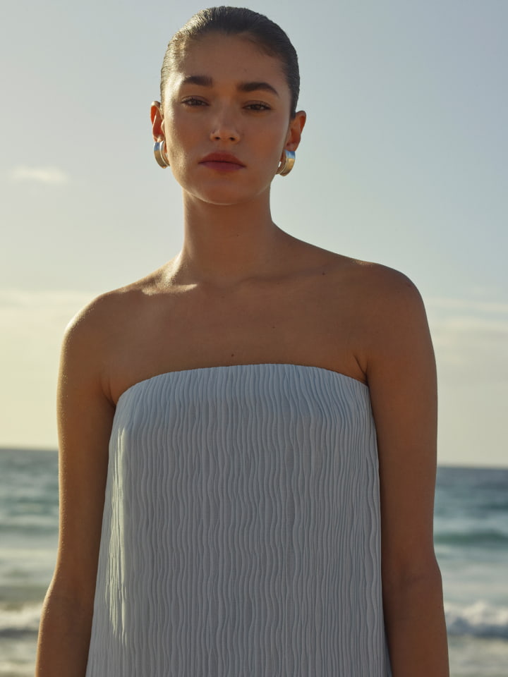 Model wearing a light blue strapless dress and silver earrings against an ocean background