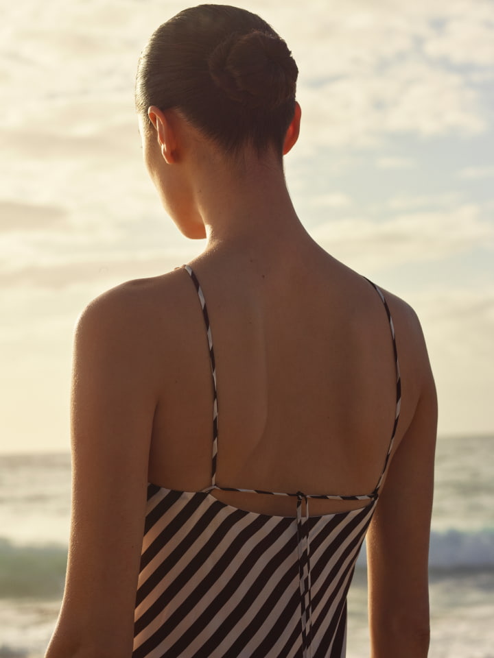 Model seen from behind wearing a black and white striped dress against an ocean and sunset sky background.