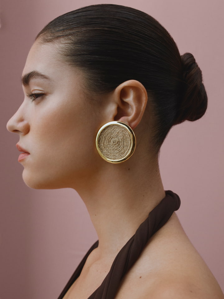 Profile shot of an adult female wearing large gold earrings with a blush-colored background, and a brown top.