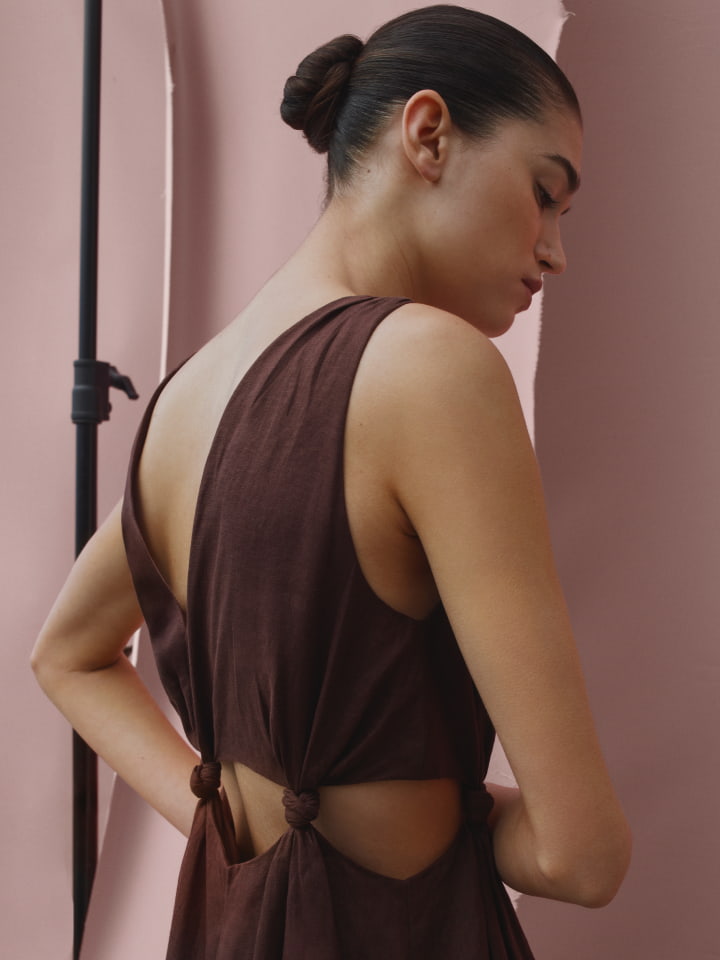 A woman with a bun hairstyle poses in a backless brown dress with tied details in a photo studio with a pink backdrop and studio lighting.