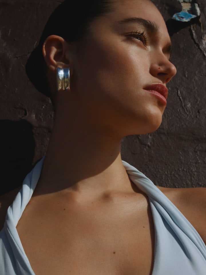 Close-up of a young adult female model with silver earrings, fair skin, and light-blue top against textured wall in natural light.