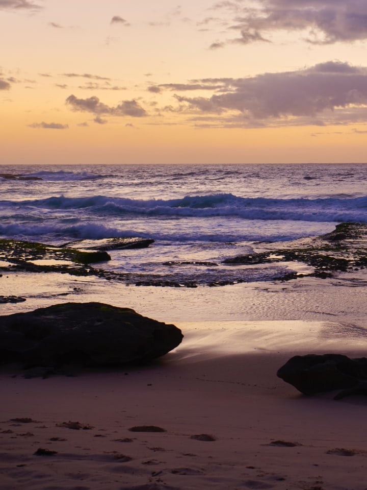 Seascape with waves rolling onto a sandy beach under a pastel sky at dusk, featuring dark rock formations and subtle cloud cover.