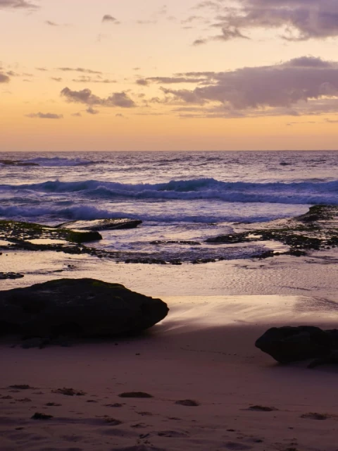 Seascape with waves rolling onto a sandy beach under a pastel sky at dusk, featuring dark rock formations and subtle cloud cover.