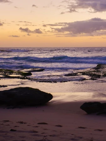 Seascape with waves rolling onto a sandy beach under a pastel sky at dusk, featuring dark rock formations and subtle cloud cover.