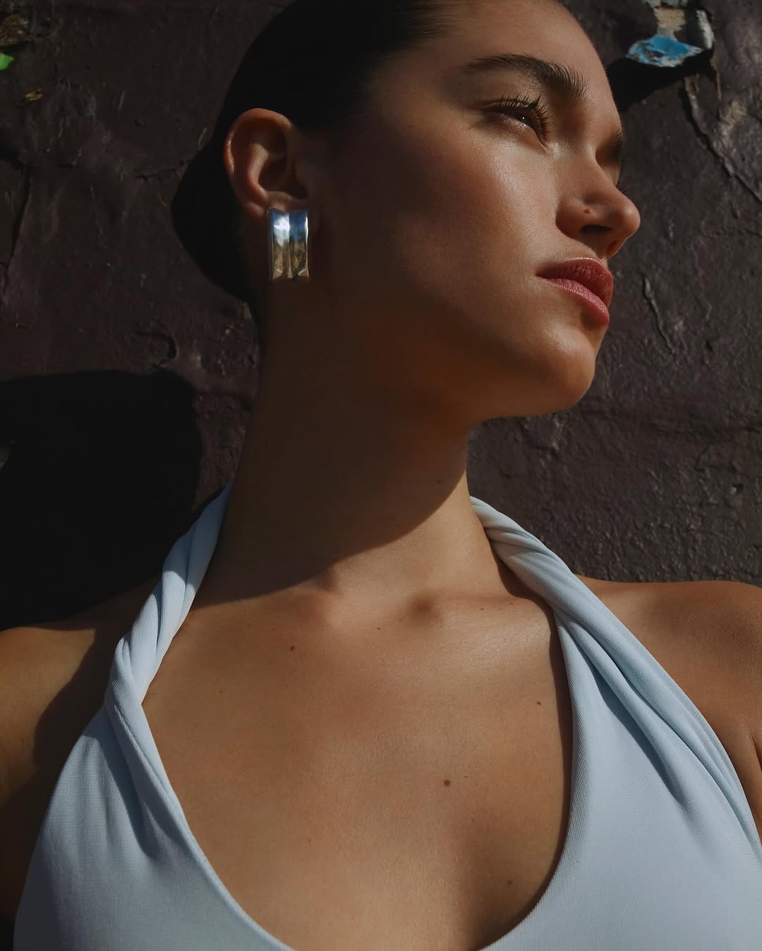 Portrait of woman with silver earrings wearing light blue top, captured in sunlight against a dark, textured background.