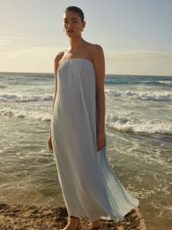 Model standing on a coastal cliff wearing a light blue strapless pleated maxi dress against an ocean backdrop.