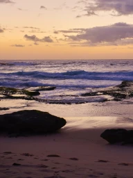 Rocky shoreline at sunset with gentle waves washing onto a sandy beach under a colorful dusk sky.