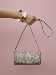 Hand holding a cylindrical snakeskin handbag against a plain pink background, wearing large gold cuff bracelets.