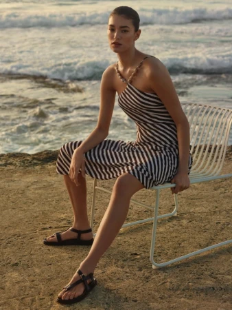 Model wearing a striped dress seated on a white wire chair by the ocean
