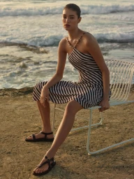 Model wearing a striped dress seated on a white wire chair by the ocean