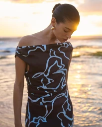 Model wearing dark patterned dress with gold earrings standing near ocean at sunset