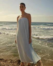 Model standing on a rocky shoreline wearing a strapless blue pleated dress against the ocean.