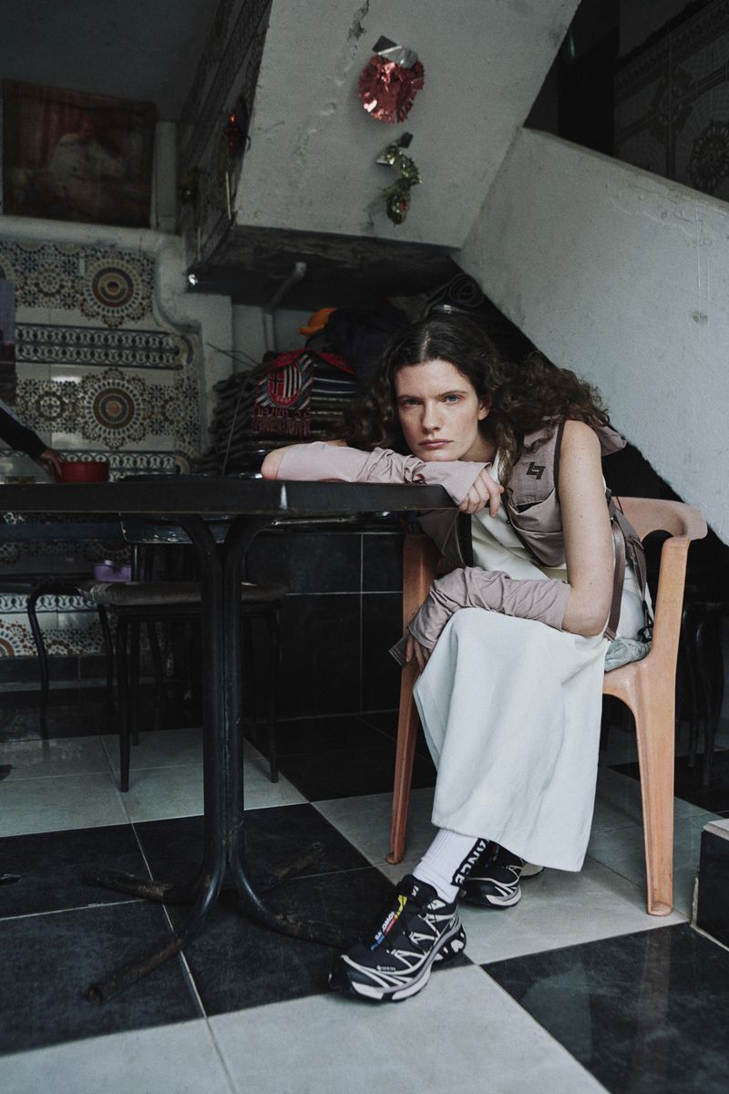 Model seated at a table indoors, wearing layered clothing and sneakers in a domestic interior with checkered tiled floors.