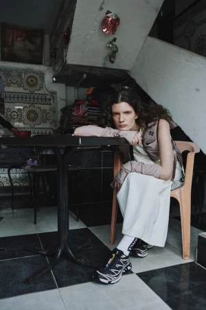 Model seated at a table indoors, wearing layered clothing and sneakers in a domestic interior with checkered tiled floors.
