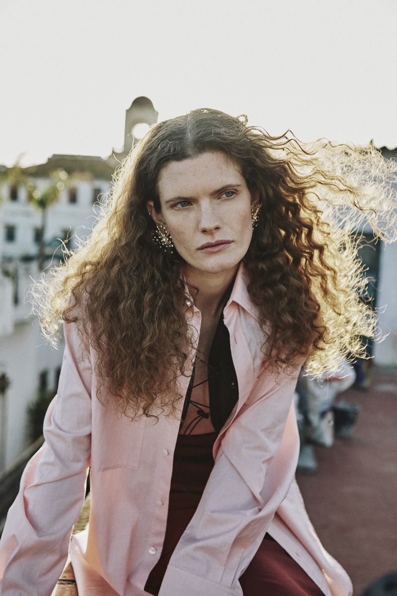 Model with long curly brown hair wearing a pink button-down shirt outdoors in an urban setting for Bare Walls.