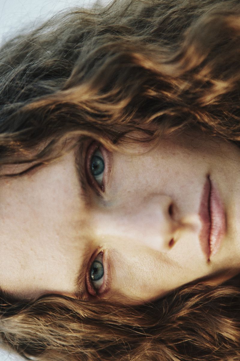 Close-up portrait of a woman with long wavy brown hair and blue eyes framed by soft natural light for The Collector Magazine.