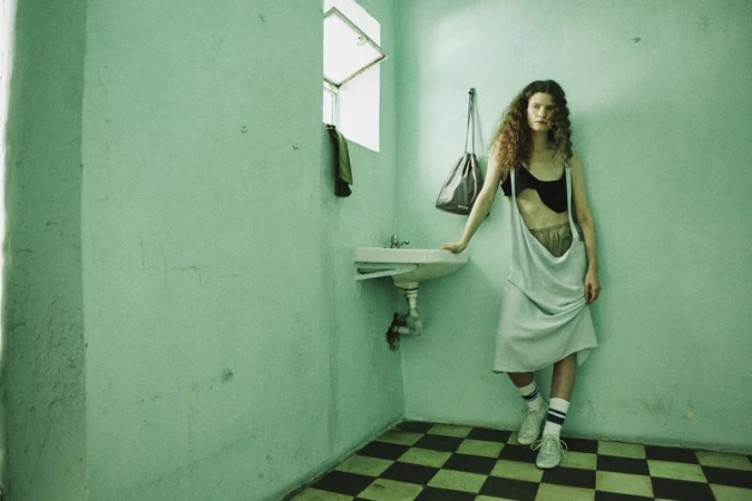 Woman with long curly hair leaning against a turquoise wall beside a sink, wearing a light blue dress over a black top and sneakers
