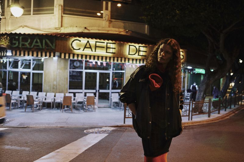 Model standing on a street at night outside a glowing cafe storefront wearing a dark jacket
