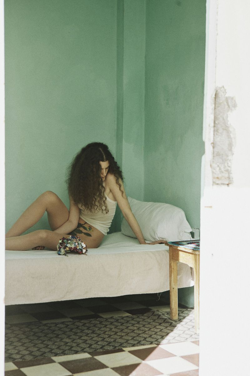 Woman with long curly hair seated on a bed in a room with green walls
