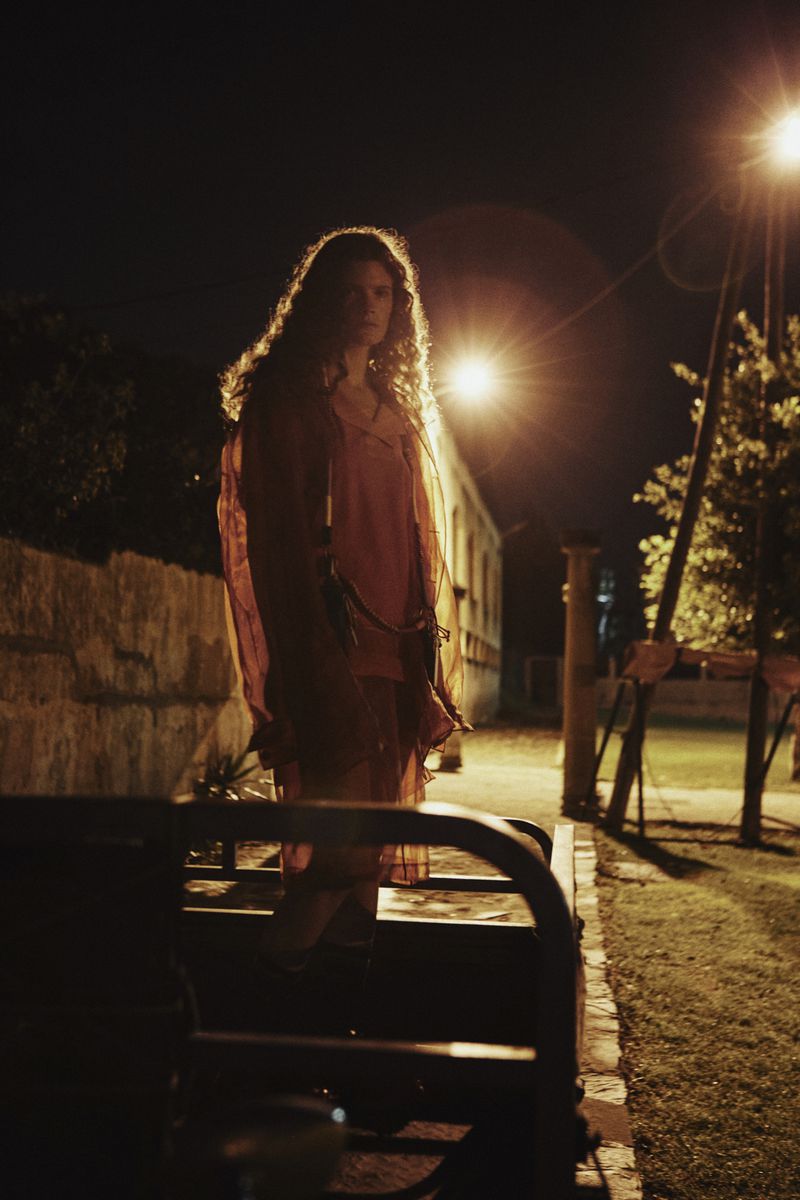 Woman with long curly hair standing outdoors at night under streetlights wearing a sheer red dress.
