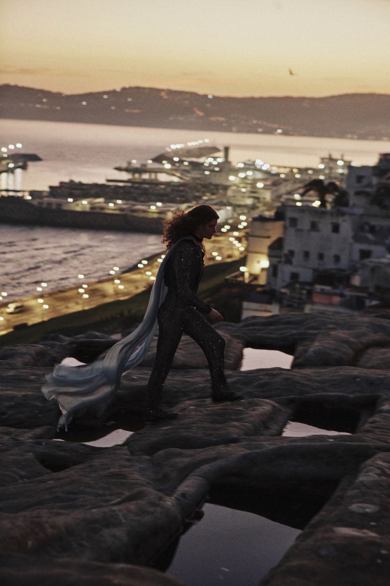 Model wearing a dark sequined jumpsuit and flowing cape walking on rocky terrain overlooking a coastal city at sunset