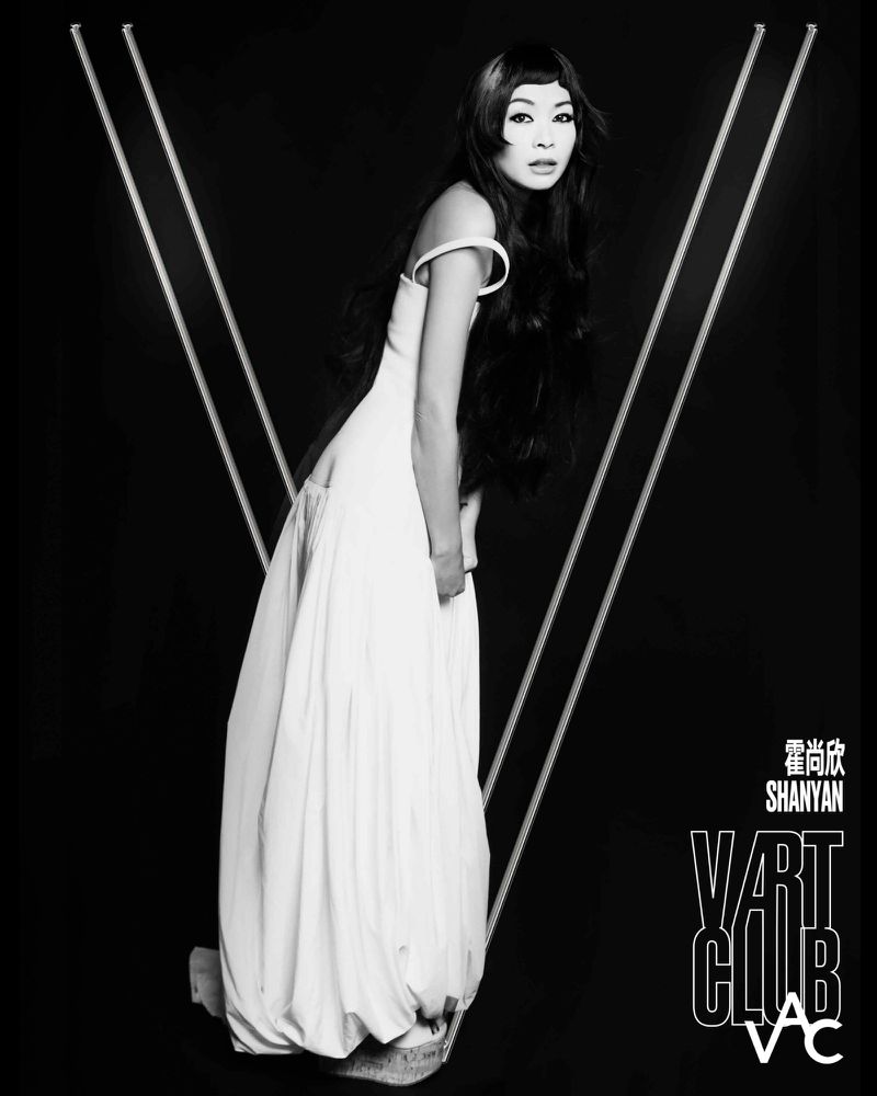 Black and white portrait of a woman in a long white gown posing inside a large letter V set against a dark background
