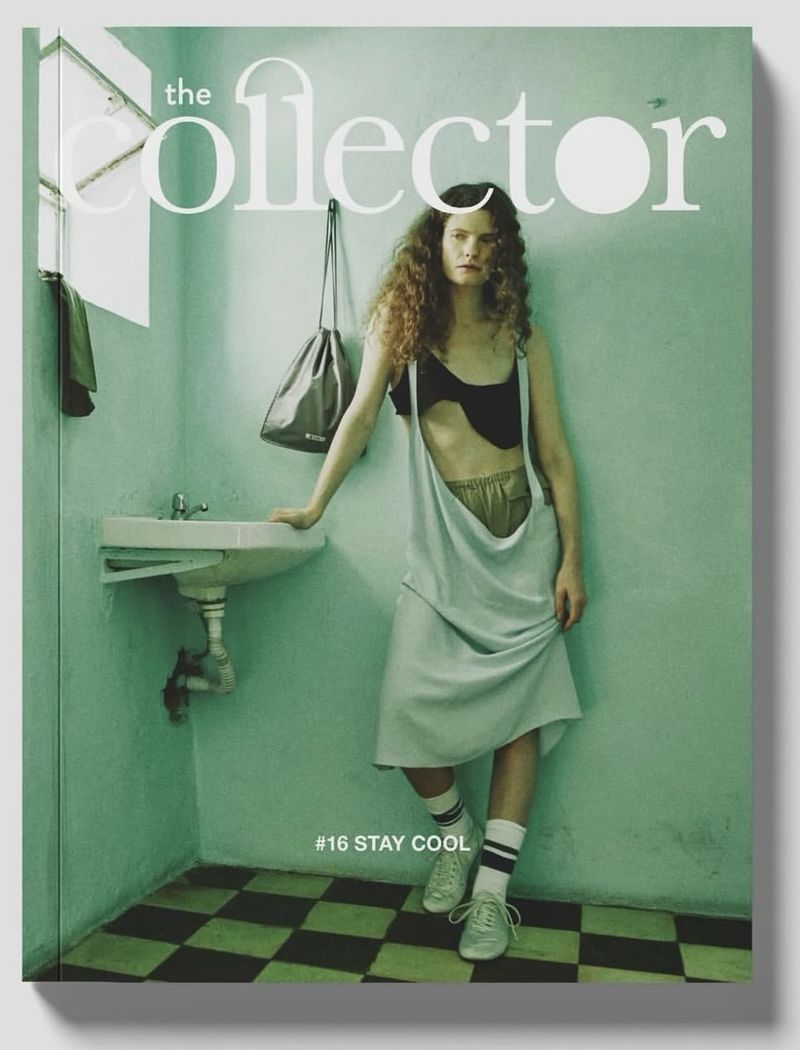 Model standing against a teal wall in a bathroom with a sink, wearing an asymmetric dress and sneakers for The Collector Magazine.