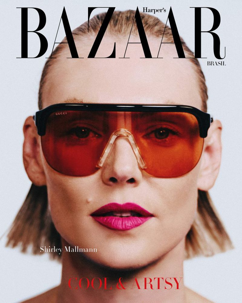 Close-up portrait of Shirley Mallmann wearing oversized amber shield sunglasses and bright magenta lipstick against a white studio background.