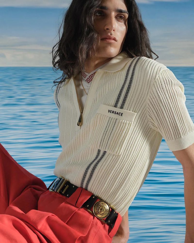 Man in cream polo shirt and coral pants poses against a blurred seascape, creating a high-fashion editorial image with warm lighting.