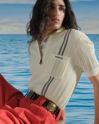 Model wearing a cream ribbed polo shirt and red trousers against an ocean background for Versace La Vacanza