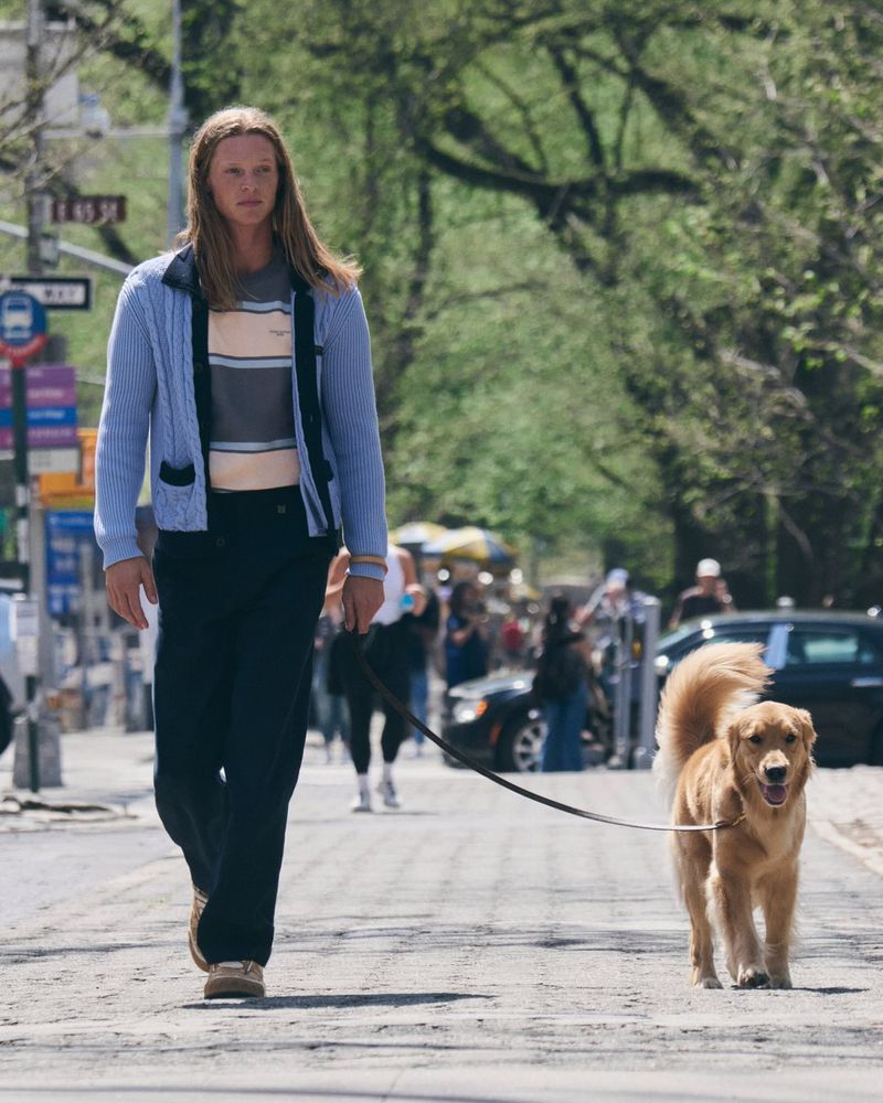 Long-haired man walking a golden retriever down a city sidewalk wearing a light blue knit cardigan and dark trousers