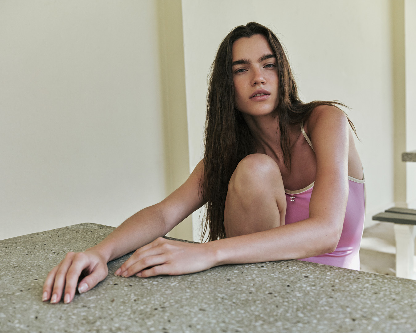 Attractive woman with long brunette hair poses in a light pink leotard, leaning on a stone table inside a minimalist indoor space.