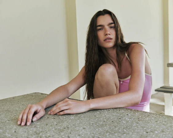 Attractive woman with long brunette hair poses in a light pink leotard, leaning on a stone table inside a minimalist indoor space.