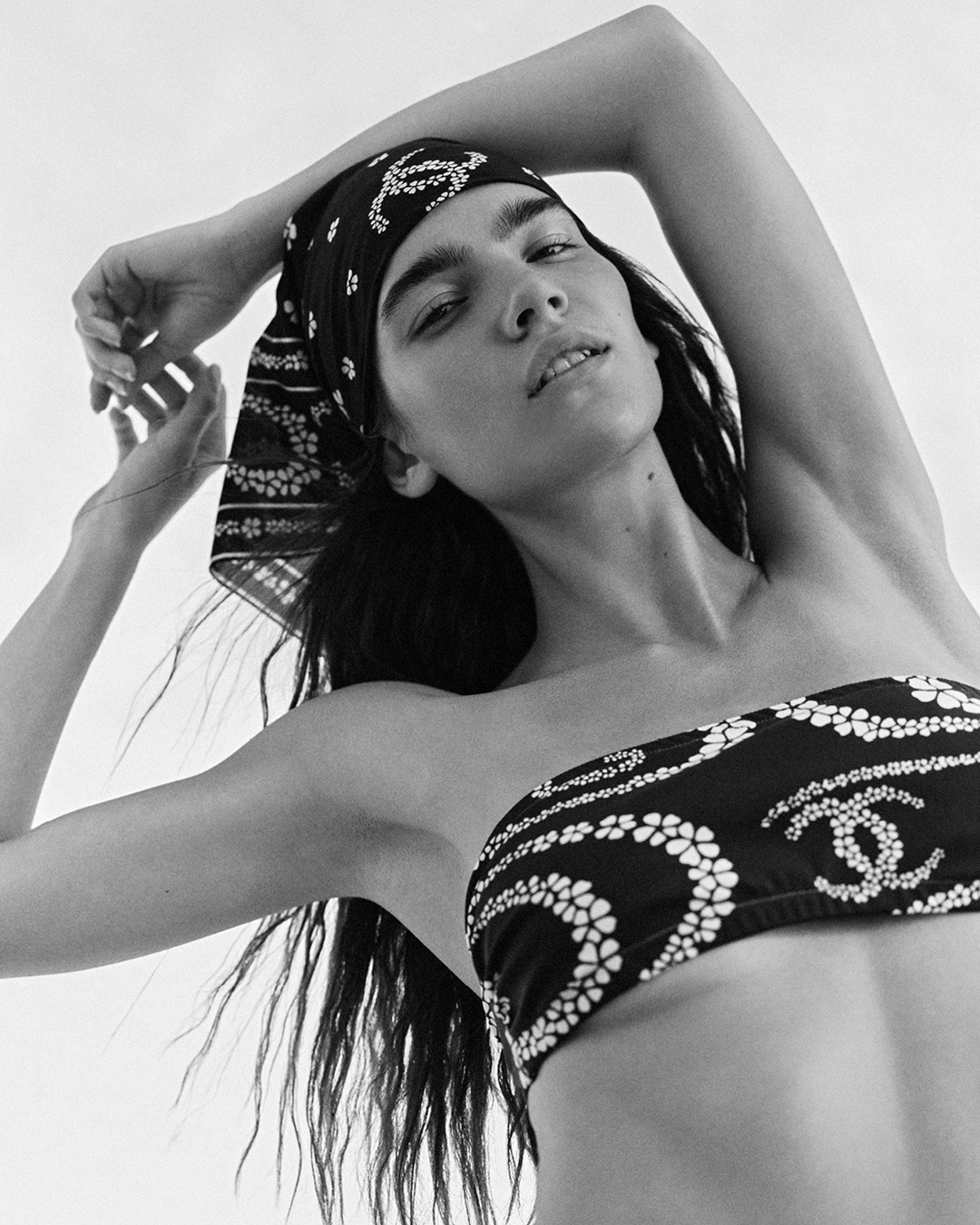 Monochrome fashion portrait of a woman wearing a printed bandeau bikini top and matching headscarf with arms raised.
