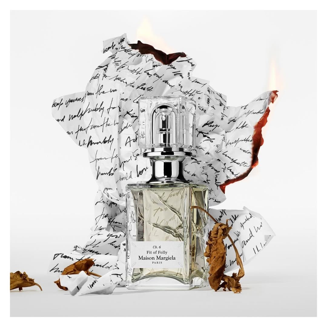 Glass perfume bottle framed by burned handwritten notes and dried leaves on a white background