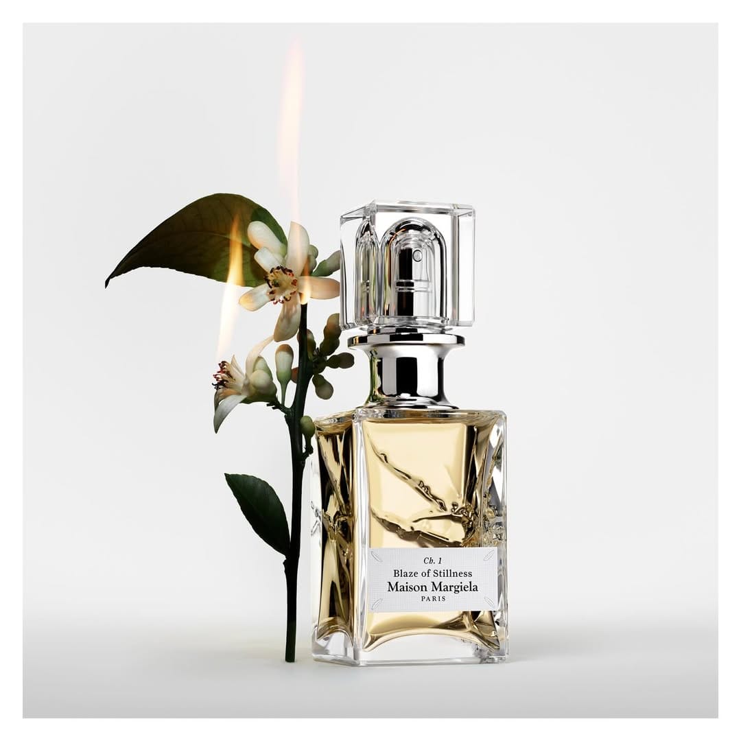 Glass perfume bottle with gold liquid next to a burning flower branch against white background