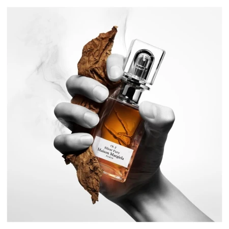 Hand holding a perfume bottle and dried leaf against a white background with delicate wisps of smoke.