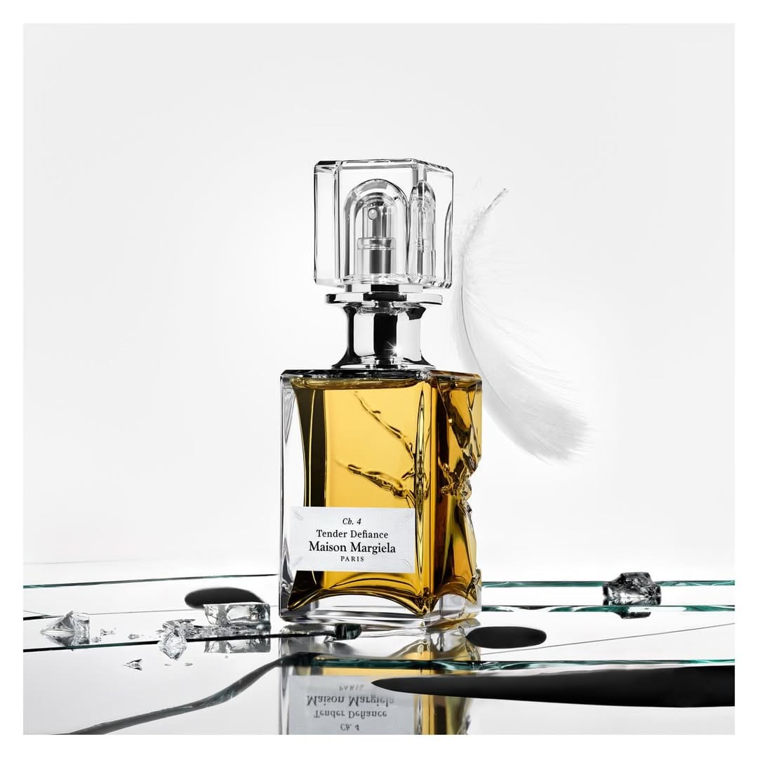 Maison Margiela perfume bottle with amber liquid on a reflective surface alongside a white feather and ice shards